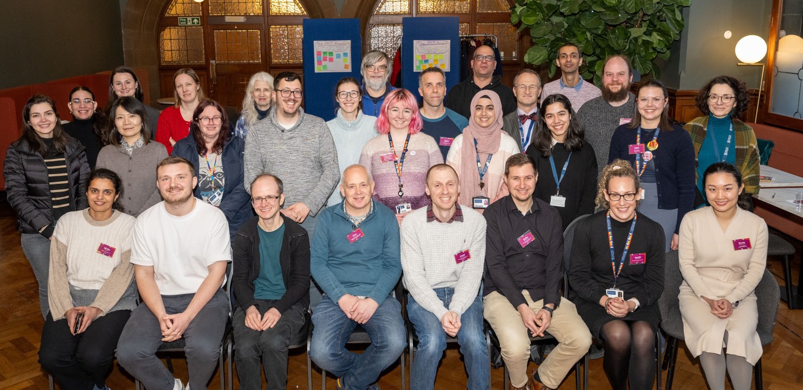 Group photo of SRTP Academy symposium attendees seated and standing in rows inside the Gilbert Scott Building, wearing conference lanyards and name badges, with event materials displayed behind them.