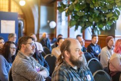 Audience members seated and listening during the SRTP Academy Symposium, wearing conference badges in a bright indoor venue with posters and greenery visible in the background.