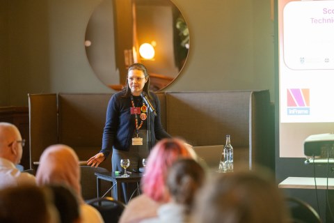 A speaker, Tabea Gerlach, presenting to an audience during the SRTP Academy Symposium, standing at a lectern with a microphone while attendees listen in a conference setting with a projected slide visible.