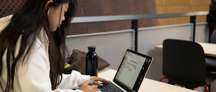 A student sitting at a desk on the first floor of the JMS building looking at their laptop, with a water bottle next to them