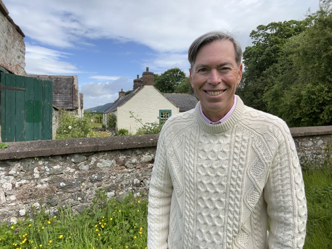 An image of a man wearing a cream jumper standing in front of an old farm buildings