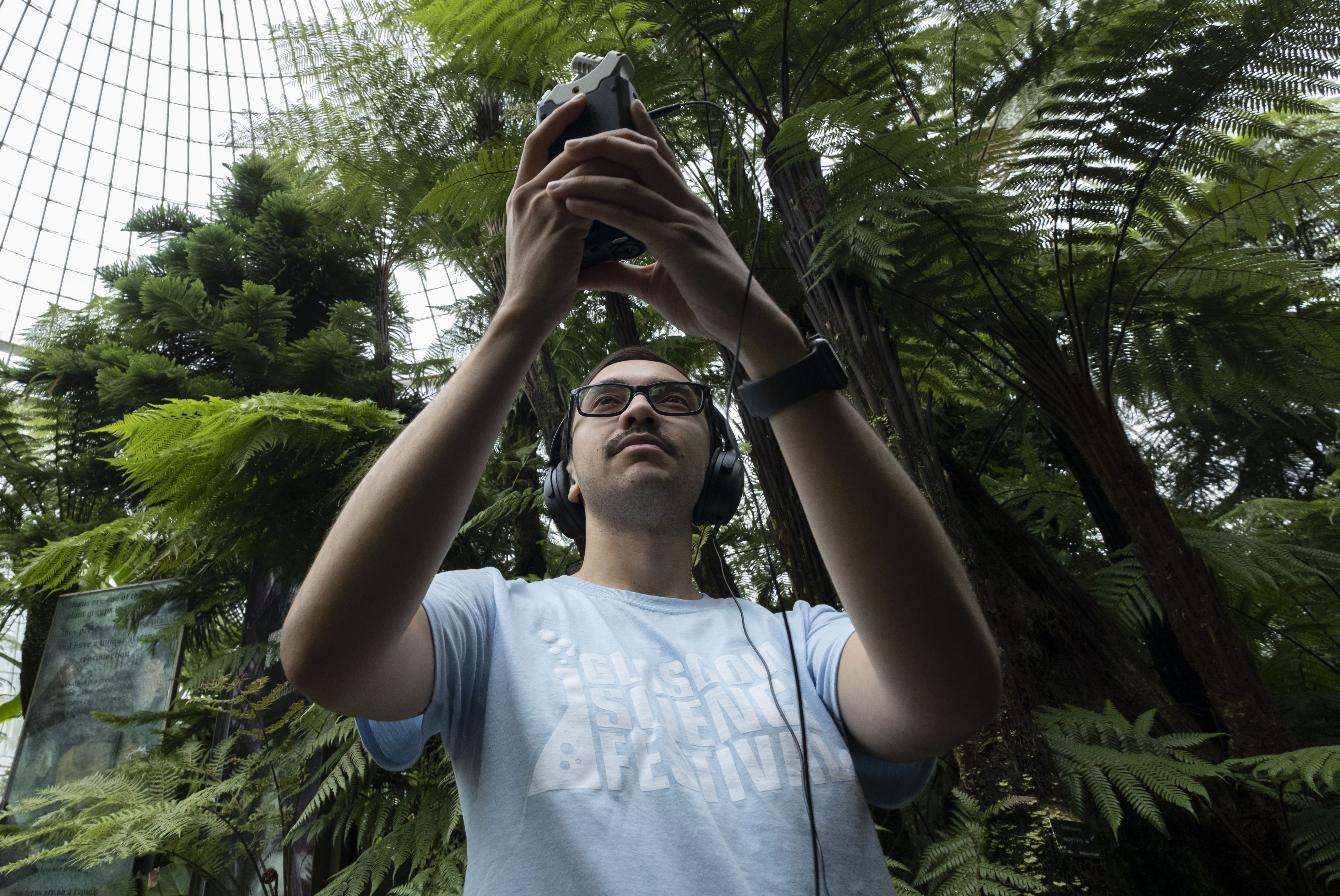 Kevin wearing a Glasgow Science Festival Tshirt holding up a small device in the Botanics Kibble palace