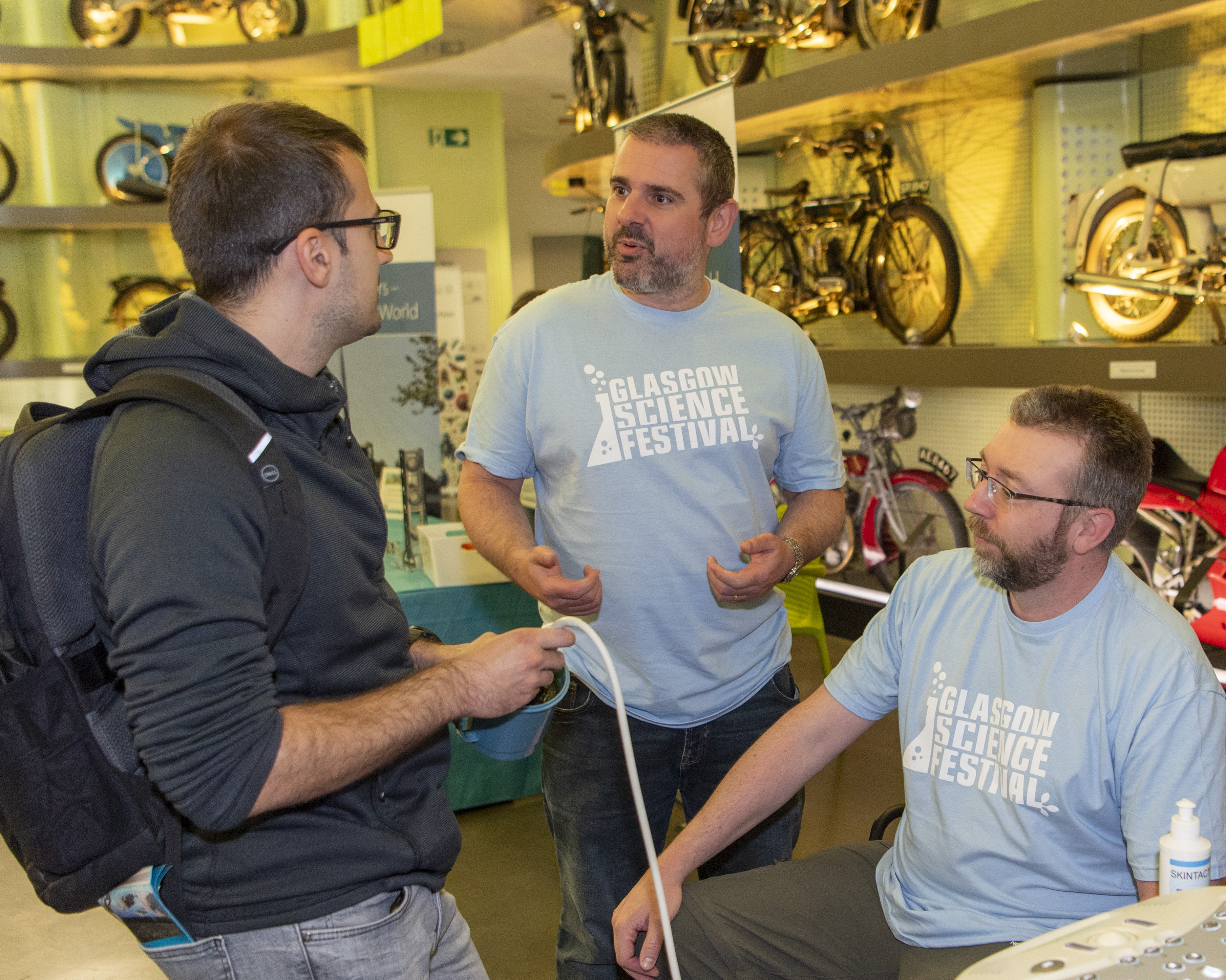 Two people wearing Glasgow Science Festival Tshirts, 1 person is holding an ultrasound probe while talking to a person. Behind is the motorbike wall in the Riverside Museum.