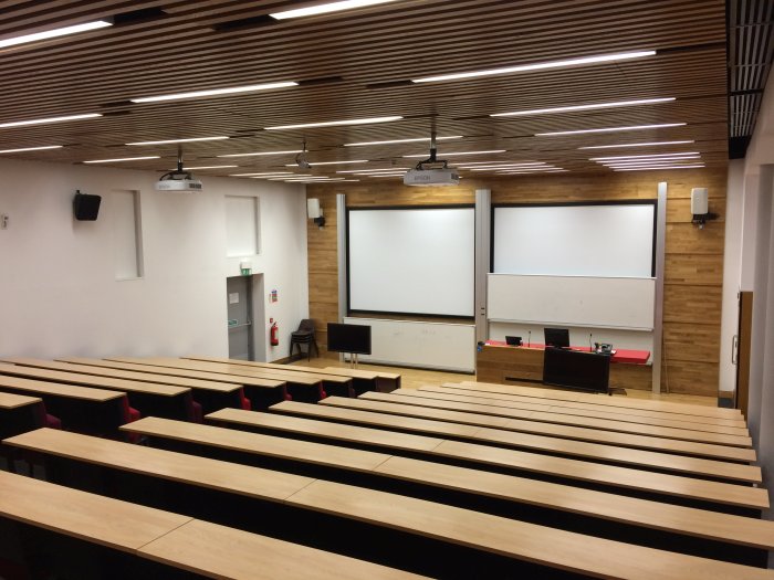 Raked lecture theatre with fixed seating, screens, whiteboards, projectors, and PC