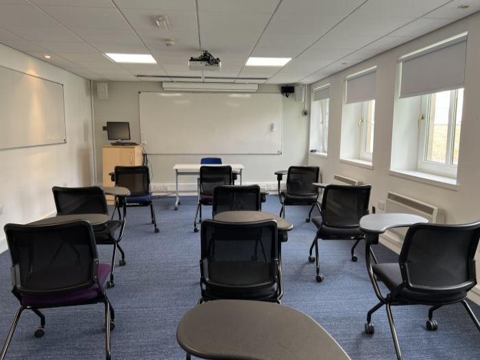 Flat floored teaching room with tablet chairs, whiteboards, projector, PC, and lecturer's table and chair.