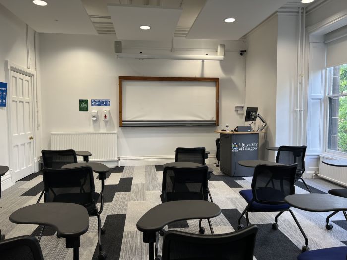 Flat floored teaching room with tables and chairs in horseshoe set-up, whiteboard, screen and PC