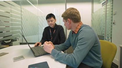 Two people sit at a small round table in a glass-walled meeting room. One person works on a laptop while the other sits beside them, leaning forward with hands clasped as they talk.