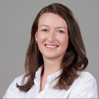 A person with long, wavy brown hair wears a white button-up shirt and is photographed against a plain grey background.