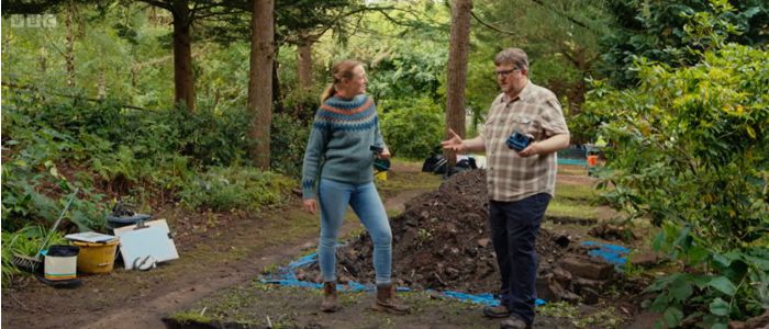 Two people standing in front of a spoilheap discuss the excavation trench before them