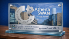 A graphic with a glass plaque sat on a wooden table, with the SGDB in the background, the Athena Swan logo on the glass, and a silver strip with the Sii logo and 'renewed until 2031' in text