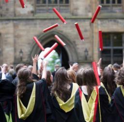 Image of a group of students wearing graduation gowns outside the University throwing their scrolls into the air