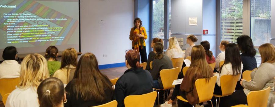 Professor Olwyn Byron stood addressing students sat in rows of chairs diagonal to her with a screen in front of them. The image is of the first Sii UG & PG Open House in 2023 and taken in the SGDB Level 2 Common Room