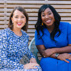 Two people are seated next to each other outdoors on woven chairs, both wearing patterned blue clothing. They are sitting close together with relaxed body posture, in front of a wooden slatted background.