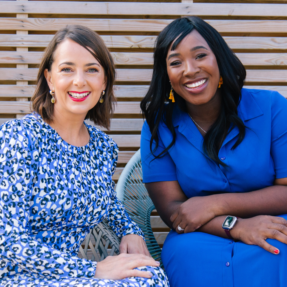 Two people are seated next to each other outdoors on woven chairs, both wearing patterned blue clothing. They are sitting close together with relaxed body posture, in front of a wooden slatted background.