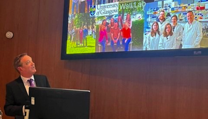 Professor Donal Wall stood at a lectern and facing a screen with images of his research group