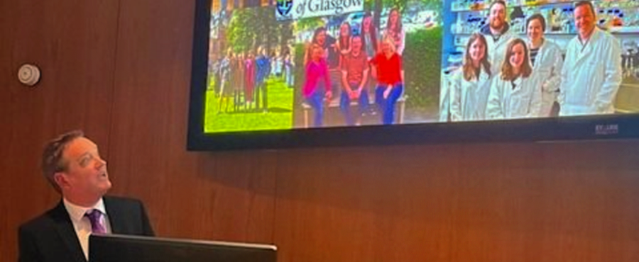 Professor Donal Wall stood at a lectern and facing a screen with images of his research group
