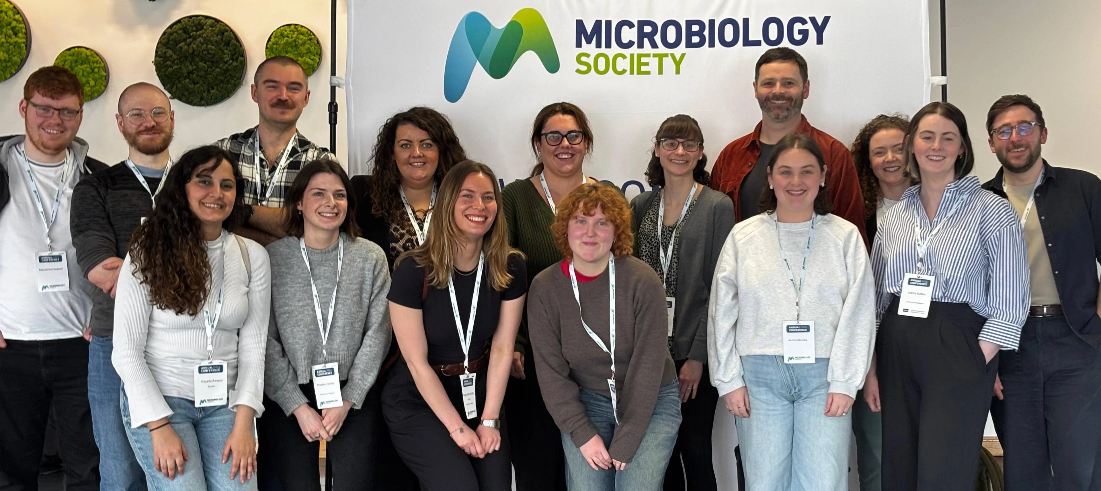A group of 15 members of the Sii community pose together indoors in front of a “Microbiology Society” banner. They are arranged in two rows, smiling at the camera, wearing casual-smart clothing and conference lanyards with name badges. The setting is a modern venue with decorative circular green wall panels behind them.