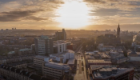 Aerial view of Glasgow city at sunset, with university and city buildings in the foreground, streets stretching into the distance, and warm golden light breaking through clouds over the skyline.