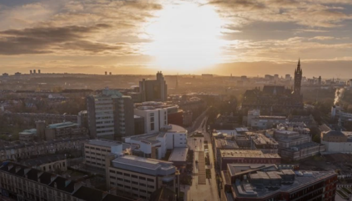 Aerial view of Glasgow city at sunset, with university and city buildings in the foreground, streets stretching into the distance, and warm golden light breaking through clouds over the skyline.