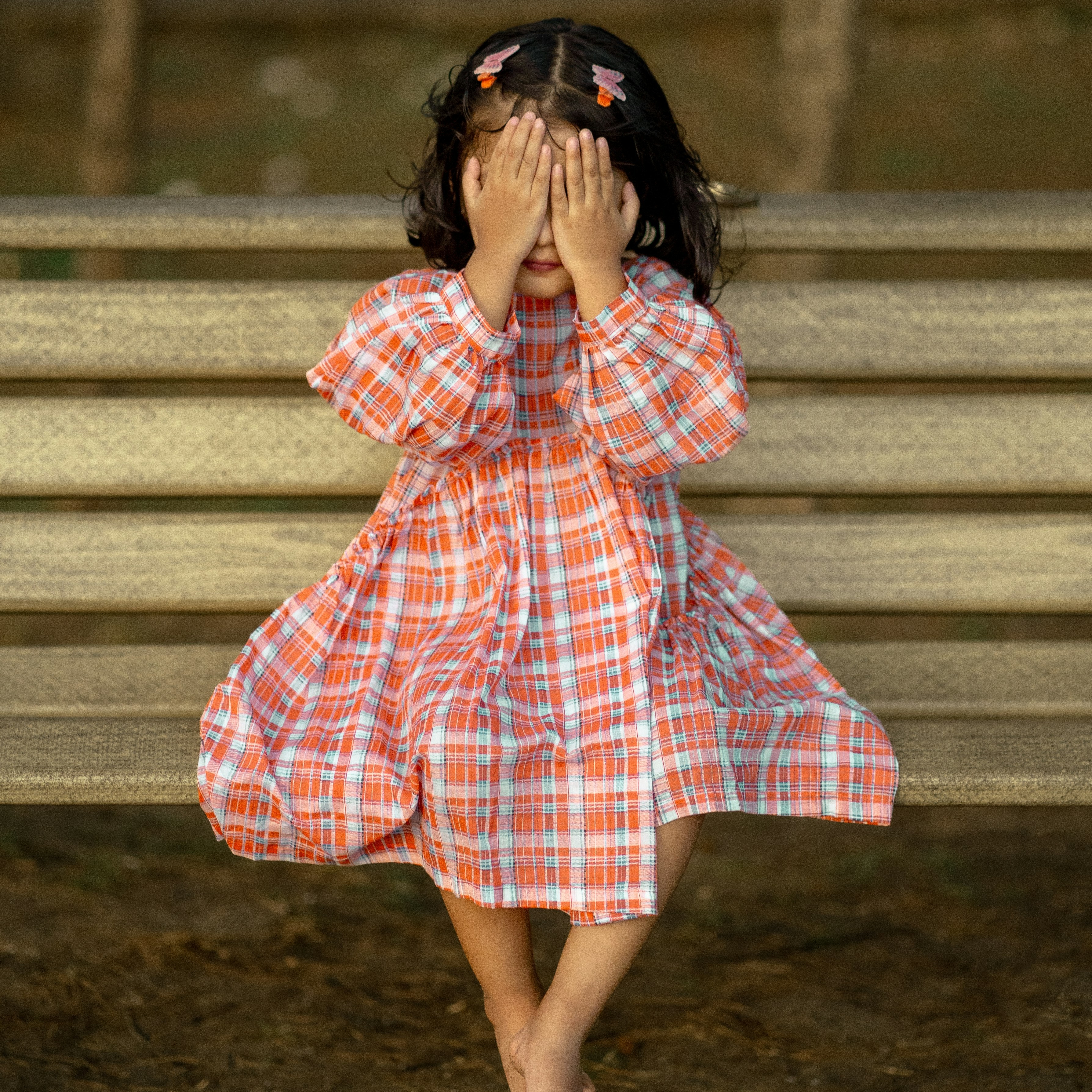 A young child sits on a wooden bench outdoors, covering their face with both hands. The child is wearing a long‑sleeved, orange and blue checkered dress, and their hair is secured with small clips. The background is softly blurred, showing the bench and ground beneath.