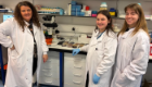 Rebecca McHugh (left) with two researchers in a laboratory, standing beside a microscope and petri dishes while wearing white lab coats.