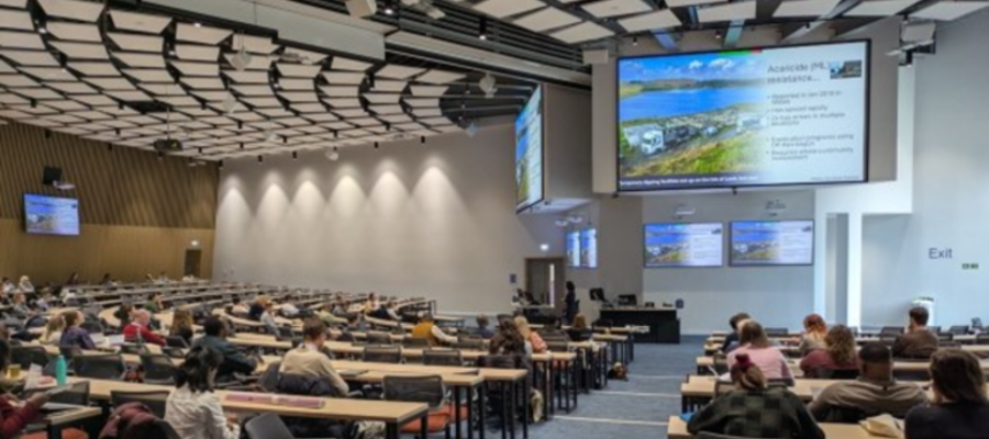 Delegates attend a presentation during the British Society for Parasitology (BSP) 2026 Spring Meeting in the James McCune Smith Learning Hub at the University of Glasgow, with researchers seated across a large lecture theatre facing multiple presentation screens