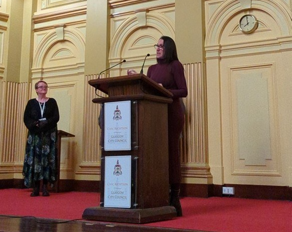 Professor Lilach Sheiner speaks at a podium during the Glasgow City Council civic reception for the British Society for Parasitology (BSP) 2026 Spring Meeting, held in an ornate civic hall with another attendee standing nearby.