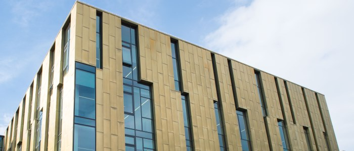 A modern multi-storey university research building with gold-toned metal cladding, tall vertical windows, and angular architectural details against a partly cloudy sky. The Sir Michael Stoker Building, home to the CVR, pictured on the University of Glasgow’s Garscube campus.