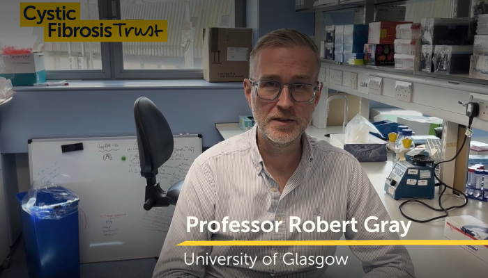 Professor Robert Gray sits in a laboratory at the University of Glasgow, facing the camera. Laboratory equipment, a whiteboard and workstations are visible in the background. The image includes the Cystic Fibrosis Trust logo and an on-screen caption identifying him as 