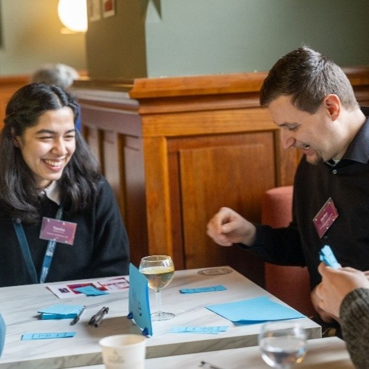 Two people sitting at a table during a workshop, working with cards and notes, with drinks on the table and name badges visible.