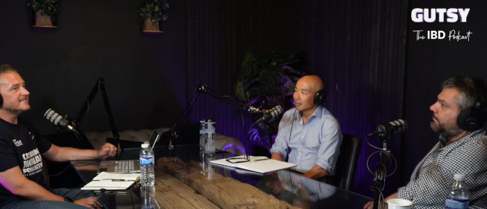 Three men wearing headphones sit around a wooden table with microphones in a podcast recording studio. Host Derek sits on the left, Professor Gwo-Tzer Ho is seated in the centre, and Professor Konstantinos Gerasimidis sits on the right. The image is from Season 2, Episode 9 of the Catherine McEwan Foundation's GUTSY podcast, where the researchers discuss IBD research and answer questions from patients and the public.