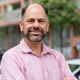 Portrait of Thomas Otto with a beard, wearing a light pink button-down shirt with his arms crossed. He is standing outdoors in front of a blurred urban background with trees and a building.