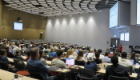 A large lecture theatre filled with researchers and students attending the inaugural Sii Research Symposium. Attendees are seated in tiered rows facing presentation screens at the front of the room, where a speaker is delivering a talk. The modern auditorium is brightly lit and nearly full, reflecting strong engagement with the event.