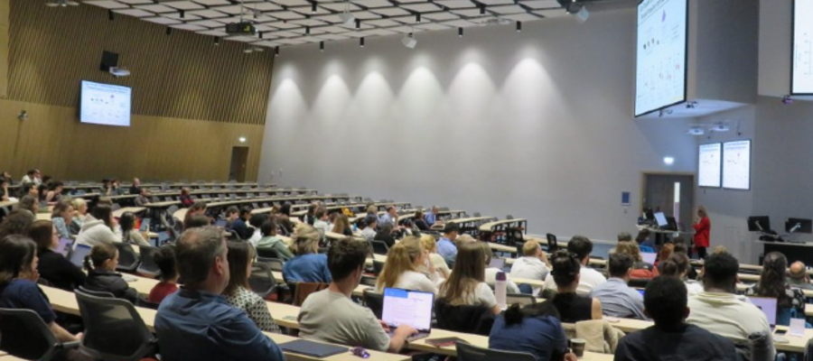 A large lecture theatre filled with researchers and students attending the inaugural Sii Research Symposium. Attendees are seated in tiered rows facing presentation screens at the front of the room, where a speaker is delivering a talk. The modern auditorium is brightly lit and nearly full, reflecting strong engagement with the event.