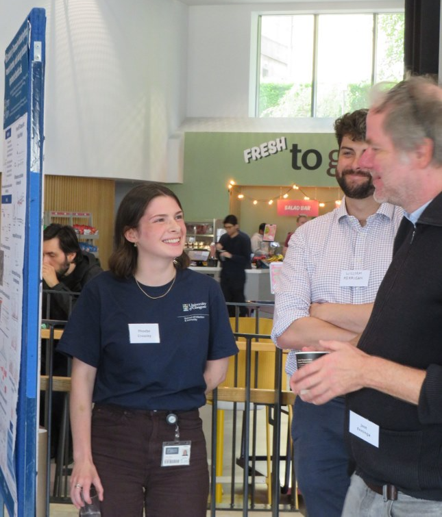 Three attendees chat beside a research poster during a poster session at the inaugural Sii Research Symposium. A presenter stands smiling next to the poster while discussing the research with two fellow attendees. The informal networking area includes café seating and refreshments, creating a welcoming environment for scientific discussion and collaboration.