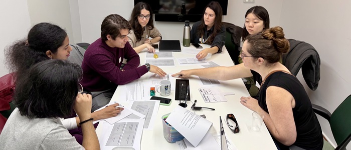 Seven students sitting around a table with pens, paper and laptops