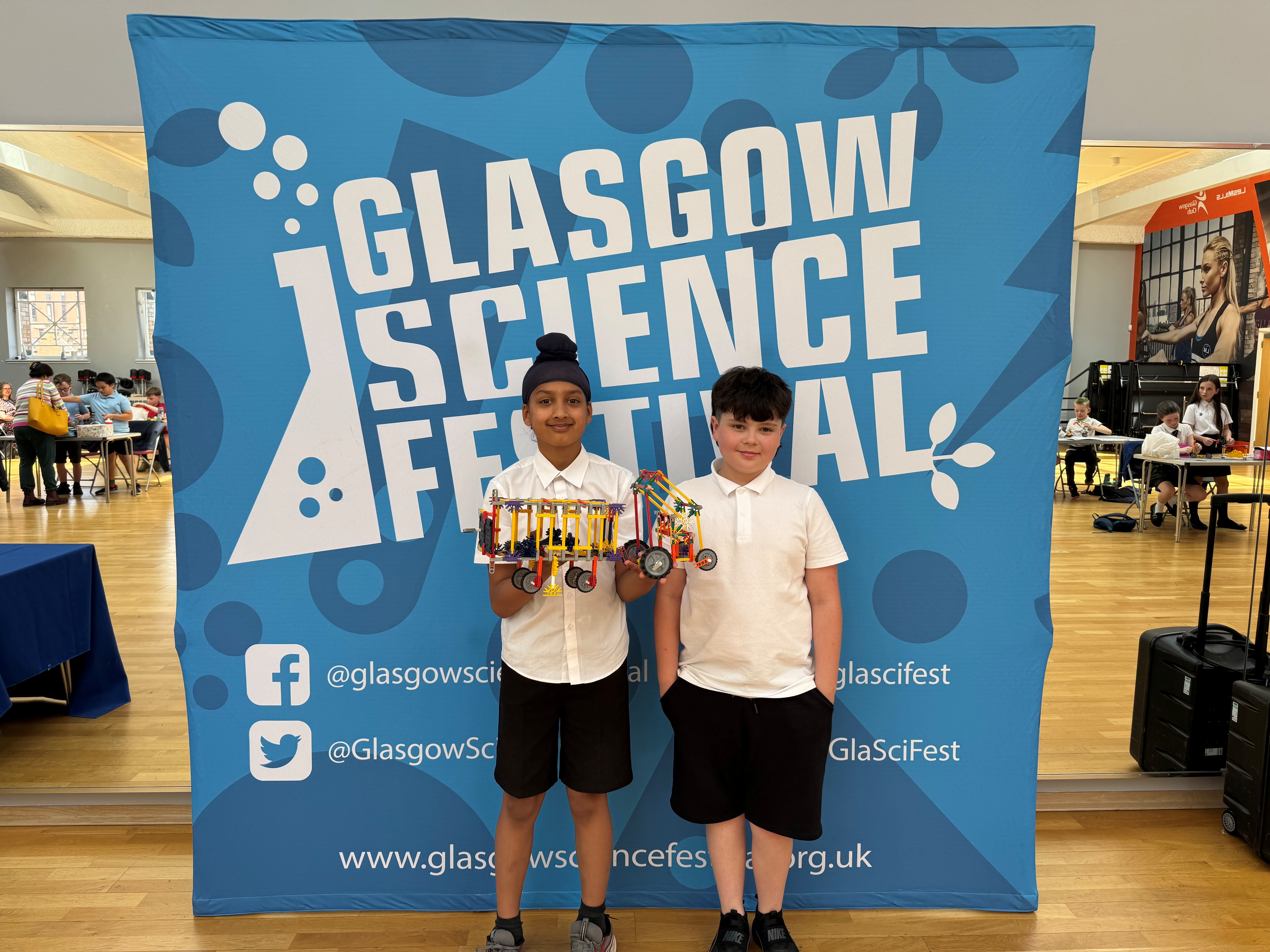 Two children holding a K'nex model standing in front of a Glasgow Science Festival banner
