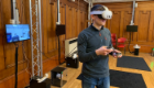 A person wearing a virtual reality headset and holding handheld controllers during a VR demonstration in a wood-panelled room, with a monitor displaying the virtual environment in the background.