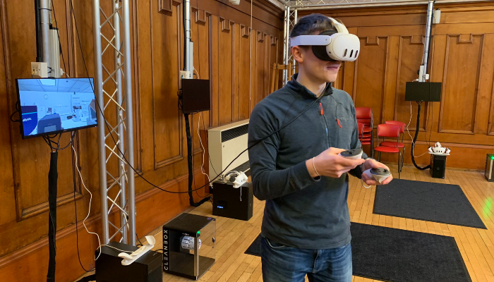 A person wearing a virtual reality headset and holding handheld controllers during a VR demonstration in a wood-panelled room, with a monitor displaying the virtual environment in the background.