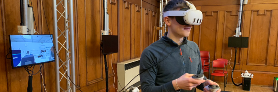 A person wearing a virtual reality headset and holding handheld controllers during a VR demonstration in a wood-panelled room, with a monitor displaying the virtual environment in the background.