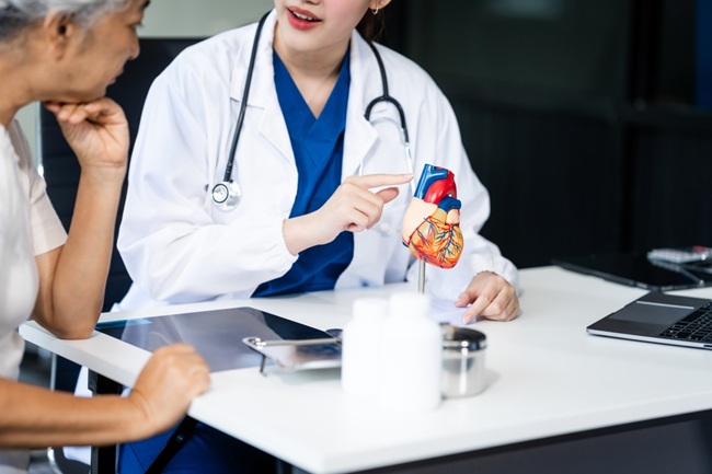 A young female nurse works at a desk with a grey-haired Asian mature woman, discussing heart-related conditions like heart disease, heart attack, and hypertension.