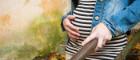 A pregnant woman wearing a stripy dress and denim jacket has a hand over her bump
