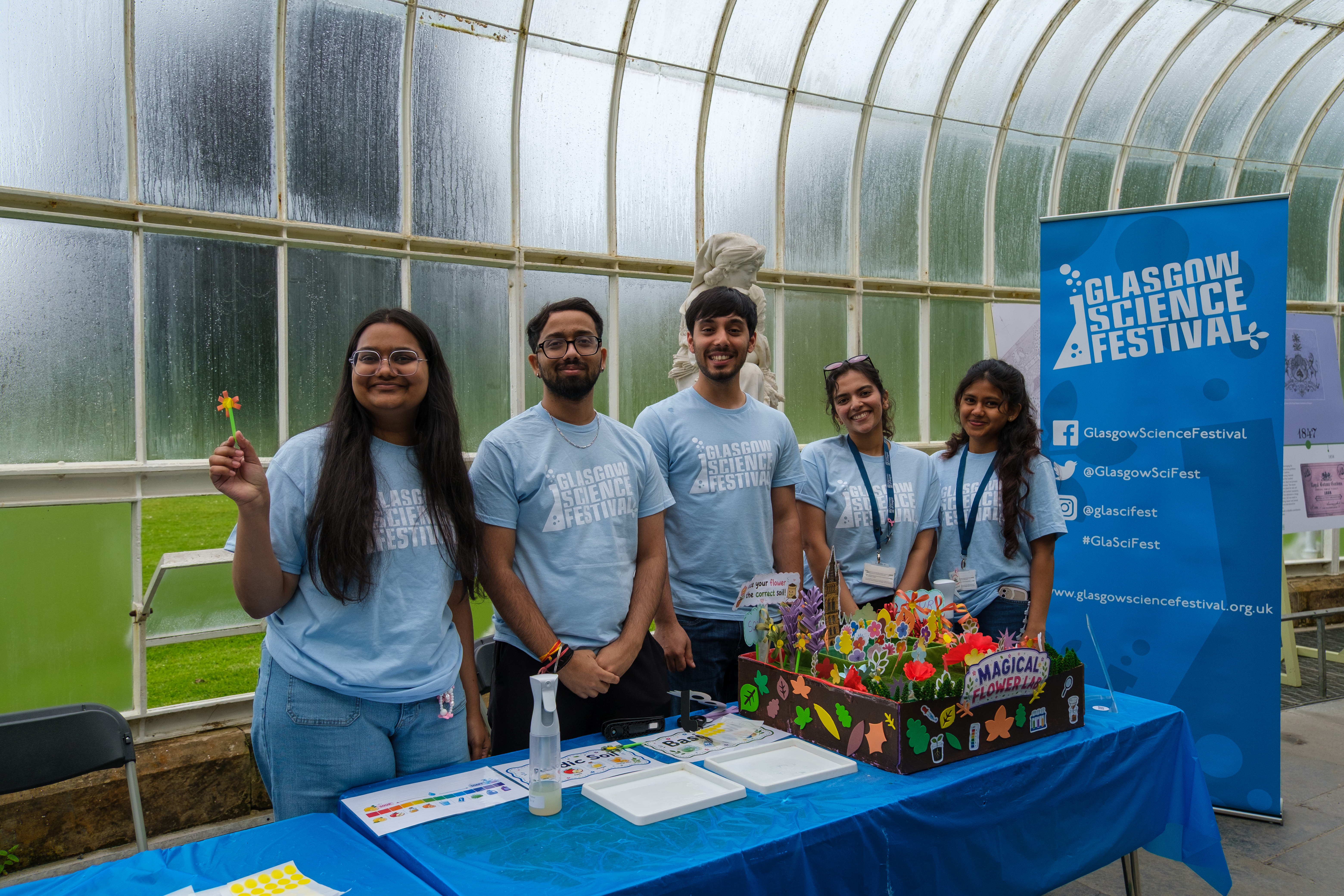 5 students standing in a glass house smiling, standing behind a table with a flower display and wearing blue Glasgow Science Festival t-shirts.