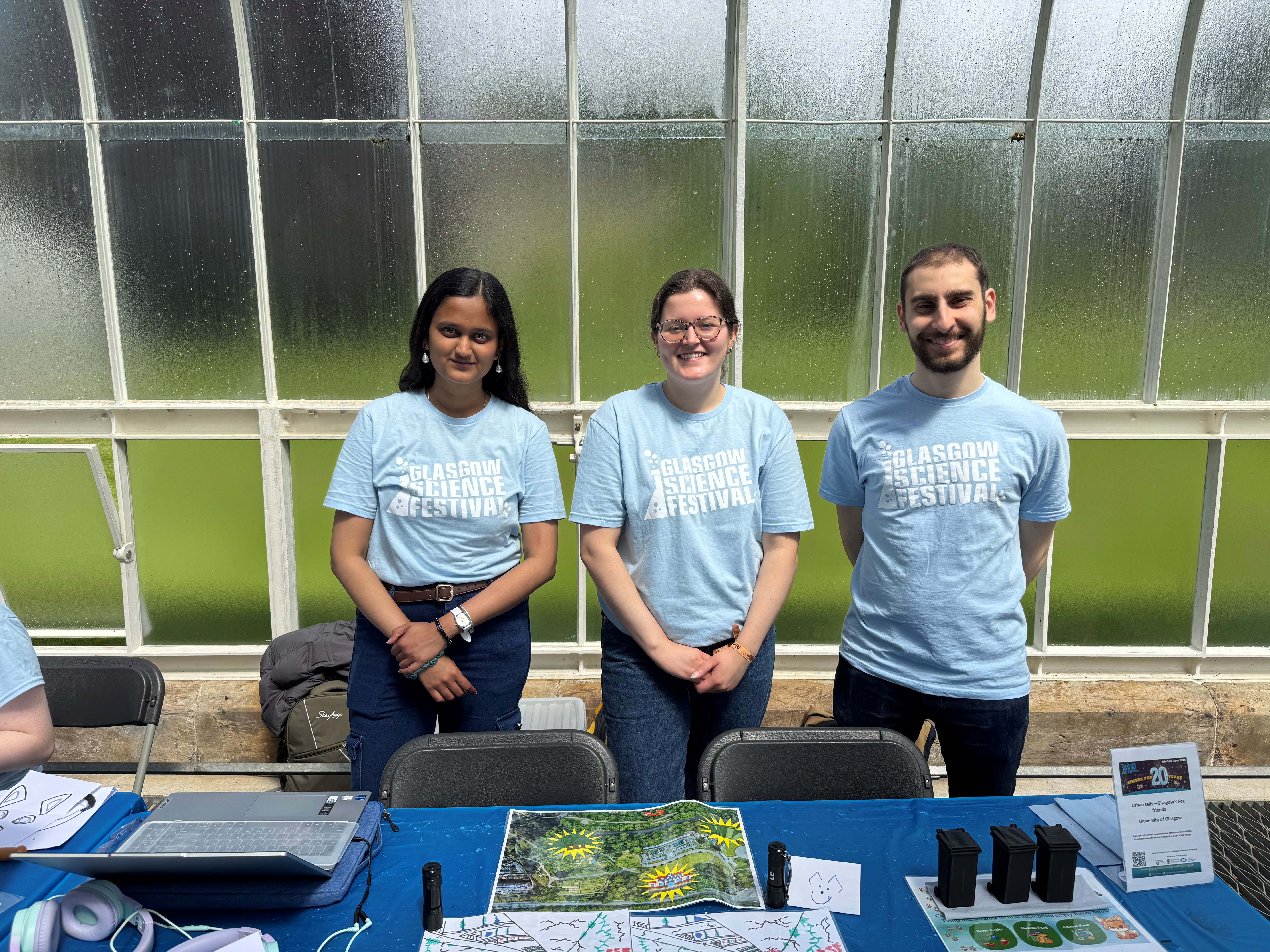 3 students standing behind a table smiling in Glasgow Science Festival t-shirts.