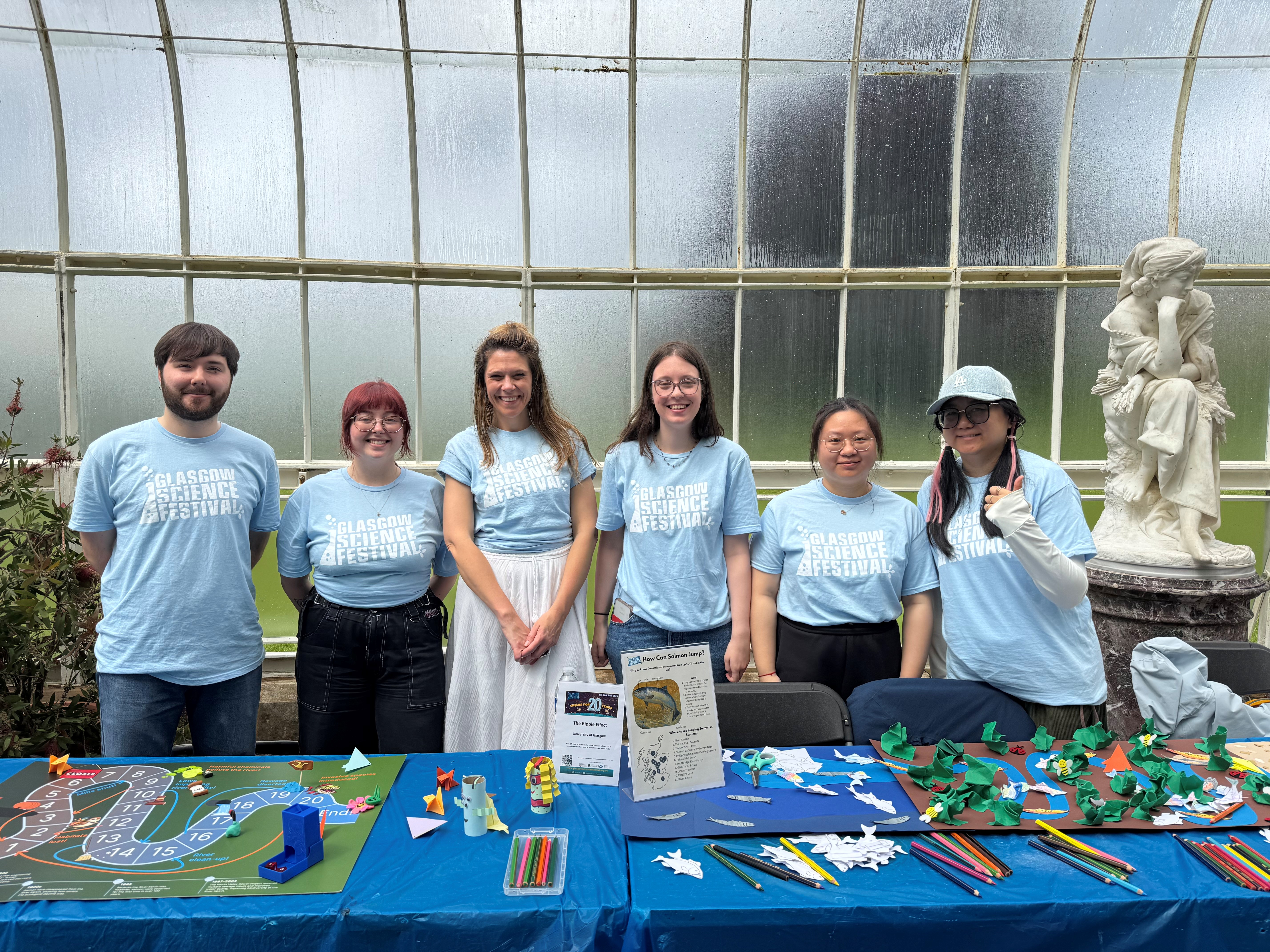 6 students standing behind a table covered in a board game and craft materials, smiling and wearing glasgow science festival t-shirts.