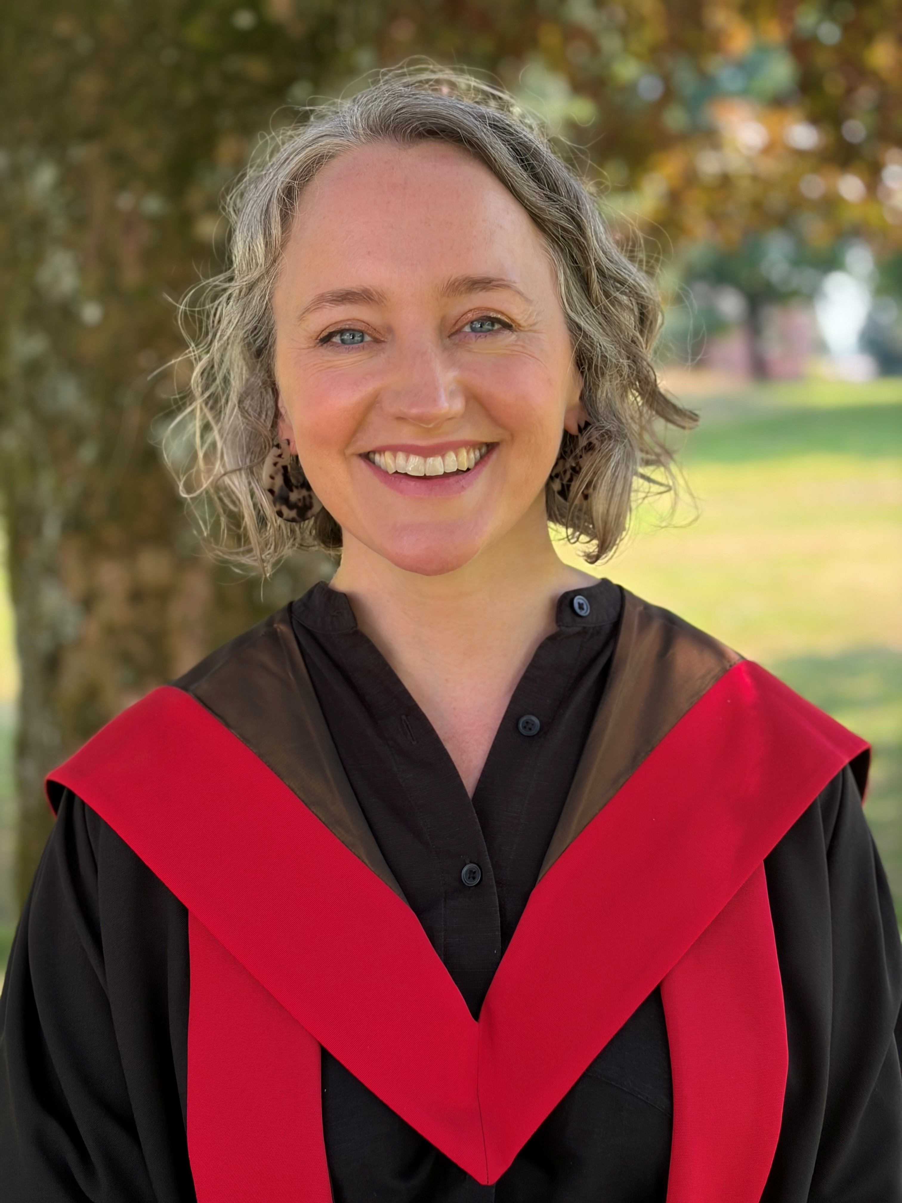 woman in graduation robes smiling in front of a tree