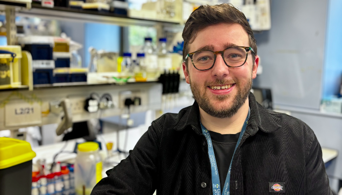Dr Liam Rooney smiles at the camera while standing in a University of Glasgow laboratory, with shelves of scientific equipment and sample bottles visible in the background.