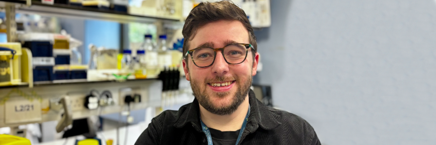 Dr Liam Rooney smiles at the camera while standing in a University of Glasgow laboratory, with shelves of scientific equipment and sample bottles visible in the background.