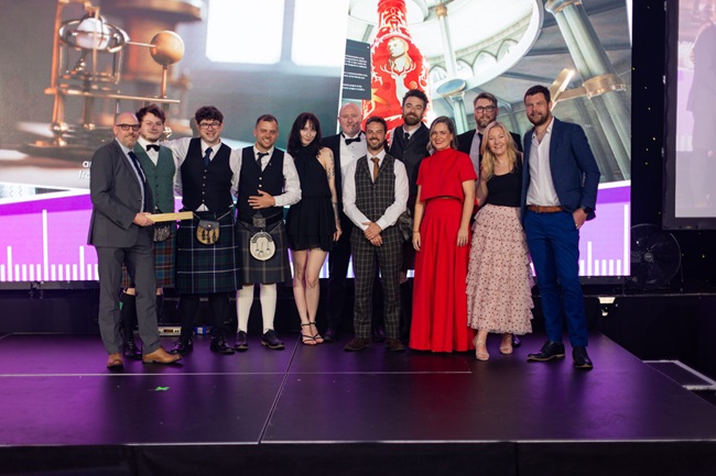 (L-R: Alastair Miller (Float); Conor Fraser (UofG); Prof Neil McDonnell (UofG); Graham Hogg (Lateral North); Agata Ostrowska (Lateral North); Andy Pennington (Float); Simon Forsythe (Lateral North); Alex McRoberts (Float): Lois Daly (Presenting Judge); Chris Harrop (Float); Prof Pauline Mackay (UofG) & Tom Smith (Lateral North) A group of people stand on a stage with a Scottish Design Award