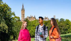 Three UofG students standing on a green with the Gilbert Scott tower and surrounding trees in the distance
